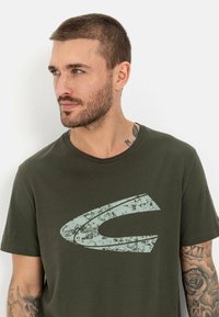 Olivenfarvet kortærmet t-shirt lavet af blød bomuld, med et slidt lyst blåt grafisk logo på forsiden.