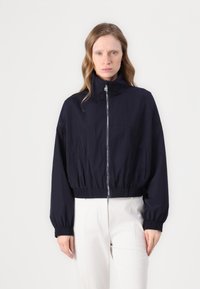 Giacca cropped navy con colletto alto, polsini elasticizzati e zip frontale. Realizzata in tessuto leggero con una texture liscia e dettagli minimi.