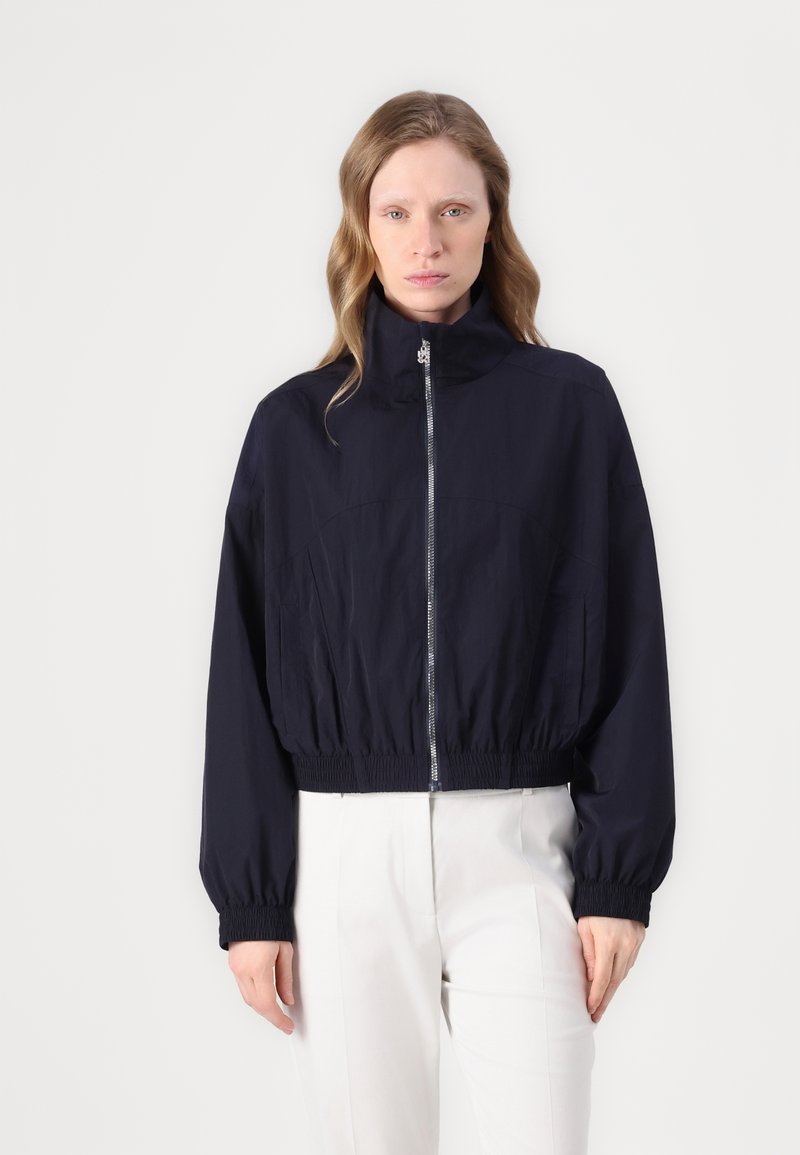 Giacca cropped navy con colletto alto, polsini elasticizzati e zip frontale. Realizzata in tessuto leggero con una texture liscia e dettagli minimi.