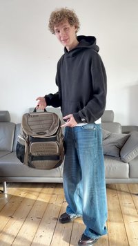 Barna és szürke hátizsák párnázott részekkel, cipzárakkal és hálós oldalzsebekkel, egy laza öltözetű személy tartja. Legfelső füllel rendelkezik.