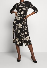 Czarna sukienka w kwiaty z trzema ćwierć rękawami, z białymi wzorami kwiatowymi, dopasowaną talią i bocznym rozcięciem, w połączeniu z czarnymi szpilkami.