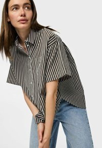 Camicia a maniche corte con colletto, caratterizzata da righe verticali marroni e bianche, tessuto morbido e vestibilità ampia, con dettaglio della tasca sulla parte anteriore.