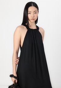 Crna halter haljina s naboranim izrezom i otvorenim stranama, izrađena od glatkog materijala. Model drži gumicu za kosu s floralnim uzorkom.