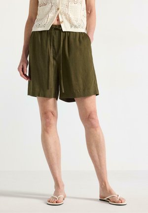 Person iført olivengrønne shorts med snøre, cremefarvet hækle-top og hvide flip-flops, stående mod en ensfarvet lys baggrund.