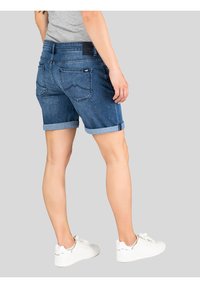Donkerblauwe denim shorts met een omgerolde zoom, twee achterzakken en een zwart label op de tailleband. Gedragen met witte sneakers.