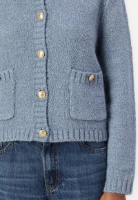 Lichtblauwe gebreide cardigan met geribbelde randen, voorzien van twee opgestikte zakken en gouden knopen. Gestyled met denim jeans.