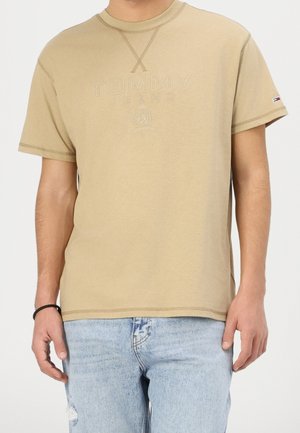 Mężczyzna ubrany w beżowy t-shirt Tommy Jeans z krótkim rękawem z subtelnym logo i jasnoniebieskie podarte jeansy na jednolitym tle.