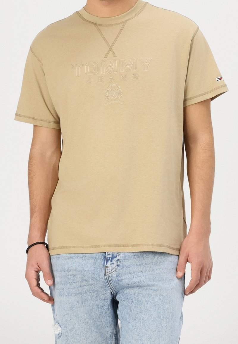 Homme portant un t-shirt beige à manches courtes Tommy Jeans avec un logo discret et un jean bleu clair déchiré sur un fond uni.