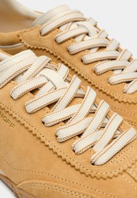 Baskets en daim beige avec des lacets crème, présentant un design festonné le long du bord et une finition texturée, avec des coutures détaillées visibles.