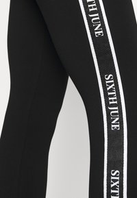 Leggings neri con striscia laterale bianca e testo "SIXTH JUNE". Materiale elasticizzato e texture liscia, progettati per comfort e flessibilità.