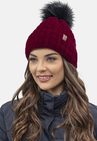 Berretto lavorato a maglia bordeaux con motivo a treccia, rifinito con un pompon in pelo sintetico nero; presenta un piccolo etichetta del marchio sul lato.