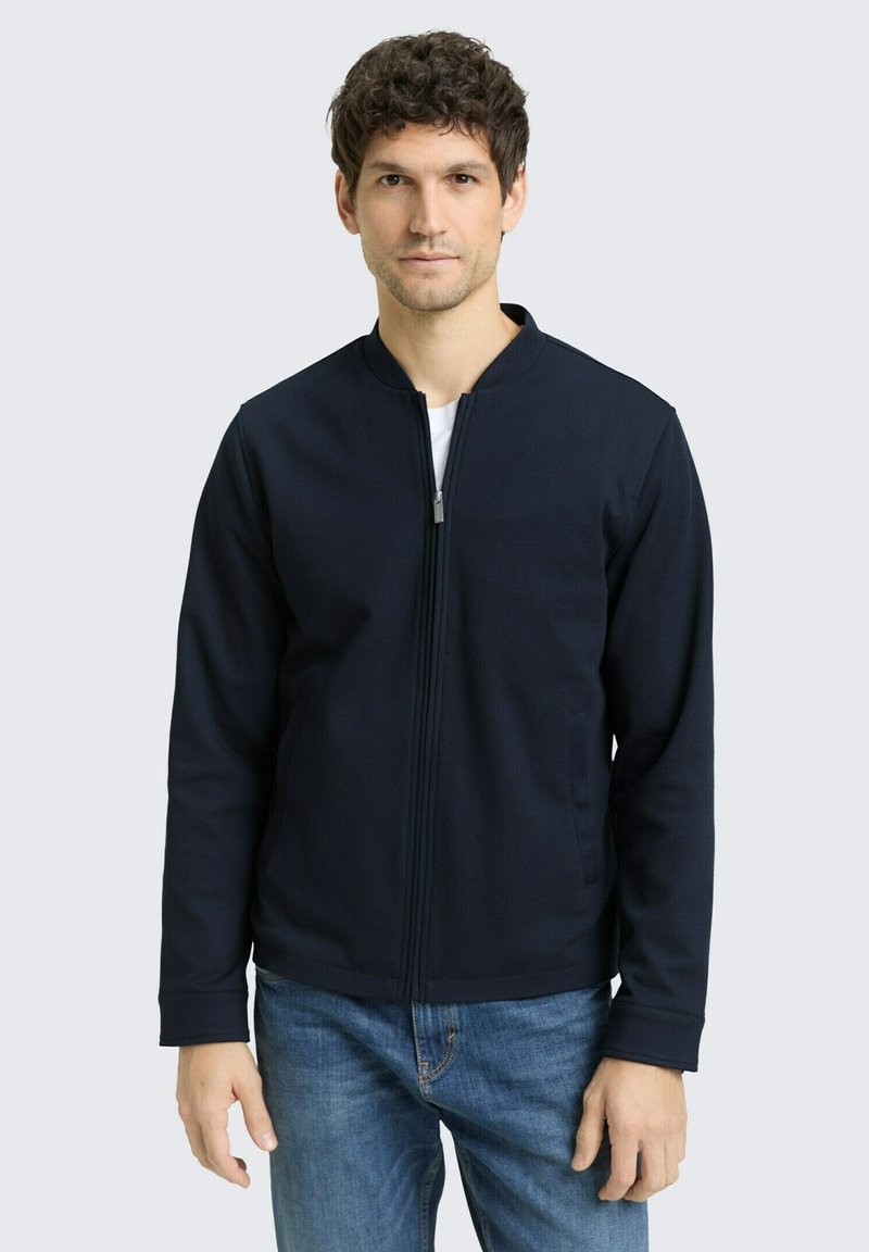Mann mit dunklem lockigem Haar, der eine dunkelblaue Jacke mit Reißverschluss über einem weißen Hemd und blauen Jeans trägt, vor einem schlichten hellen Hintergrund steht.