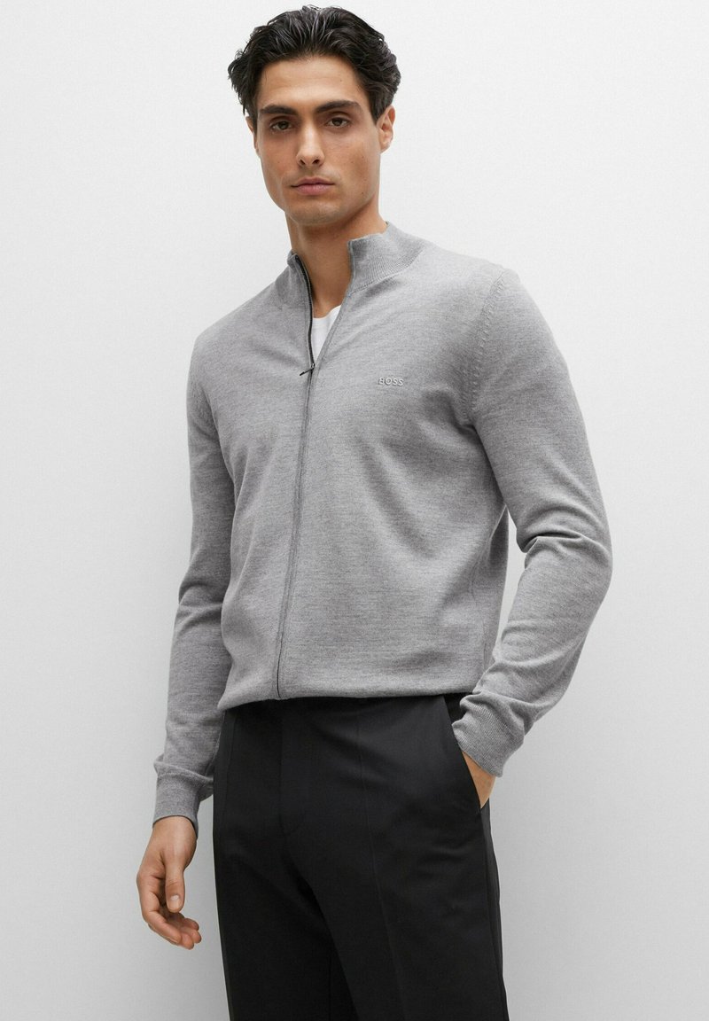 Gray zip-up sweater made of knit fabric, featuring long sleeves, a high collar, and a small brand logo on the chest. Paired with black trousers.