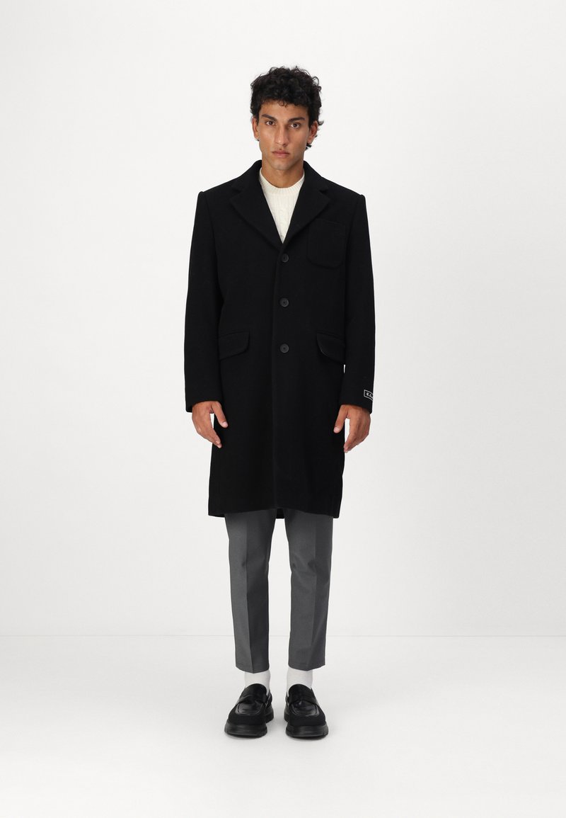 Schwarzer Wollmantel mit Reverskragen, Knopfverschluss, einer Brusttasche und zwei Seitentaschen, getragen über einem cremefarbenen Pullover und grauen Hosen.