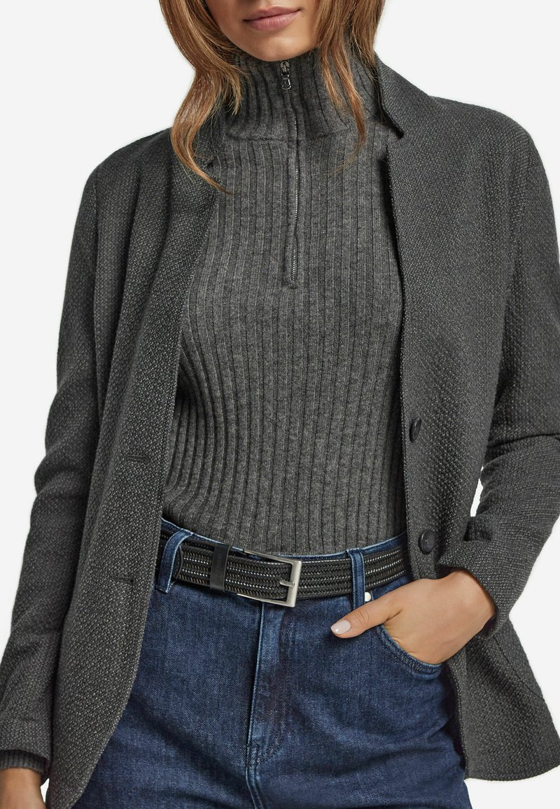 Grå strukturerad kavaj över en ribbad grå polotröja, kombinerat med höga blå denimjeans och ett svart flätat bälte med silverbuckla.