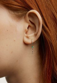 Aro de oro con una piedra marquise verde y tres gemas transparentes. Banda texturizada con un brillo sutil. Llevado en la oreja de una persona.