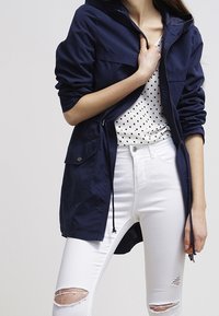 Veste légère bleu marine avec capuche, fermeture éclair et plusieurs poches avant ; portée sur un haut blanc à pois et un jean blanc déchiré.