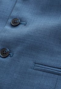 Tumed sinine tekstuuriga kangas lähivõte, millel on kaks musta nööpi ja horisontaalne õmblusdetail, mis meenutab pintsakutaskut või jopet.
