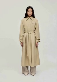 Casaco trench bege com gola e cintura cingida. Mangas longas com detalhes nos punhos. Textura suave e corte reto, combinado com calças claras.