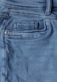 Tela de mezclilla azul claro con un bolsillo trasero, costuras visibles y un acento de botón plateado. La textura es suave y ligeramente desgastada.