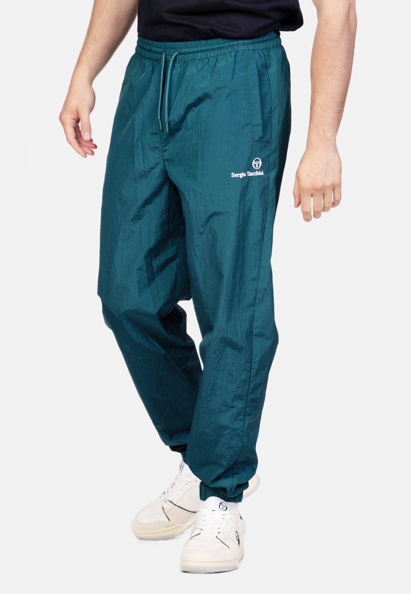 Muž v tealových teplákových nohaviciach Sergio Tacchini a bielych teniskách, stojí pred jednoduchým svetlým pozadím.