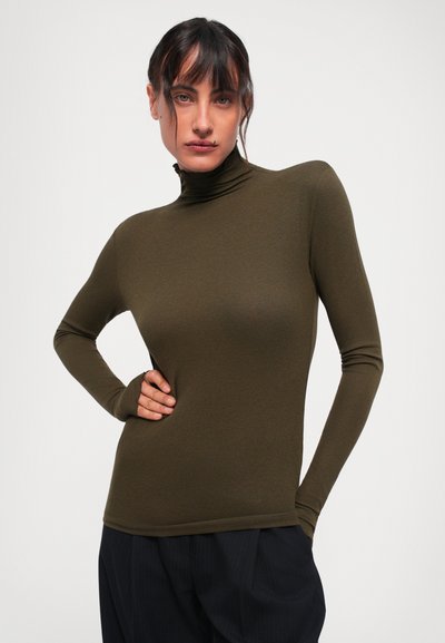 Woman wearing olive green fitted turtleneck and dark loose trousers, posing with one hand on hip against plain background.