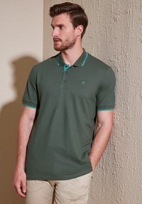 Temnozelena polo majica s kratkimi rokavi, ki se ponaša s turkiznimi detajli na ovratniku in rokavih, izdelana iz gladkega in zračnega materiala.