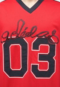 Κόκκινο αθλητικό μπλουζάκι με τον αριθμό 03 σε μαύρο και άσπρο περίγραμμα και μαύρο καλλιγραφικό κέντημα πάνω από τον αριθμό.