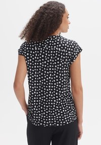 Zwarte blouse met een wit vierkant patroon, korte mouwen en een ronde zoom. Stof lijkt glad met een lichte rek.