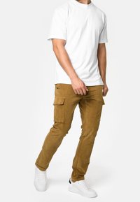 Braune Cargo-Hose mit zwei Seitentaschen, kombiniert mit einem schlichten weißen T-Shirt und weißen Sneakers. Die Hose hat eine weiche Textur und einen Slim-Fit.