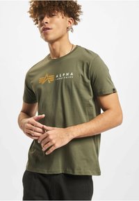 Bombažno majico kratkih rokavov olivno zelene barve z logotipom "ALPHA INDUSTRIES" v belem in zlati barvi s stilizirano ikono, mehke teksture.