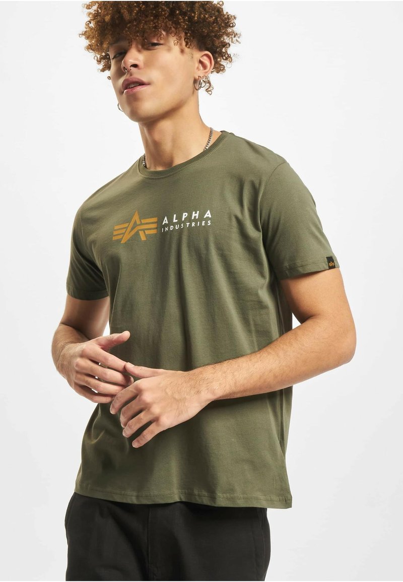 Bombažno majico kratkih rokavov olivno zelene barve z logotipom "ALPHA INDUSTRIES" v belem in zlati barvi s stilizirano ikono, mehke teksture.