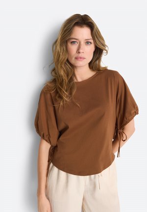 Donna con capelli castano chiaro ondulati, che indossa una maglietta marrone a maniche corte con dettagli annodati e pantaloni beige chiaro, in piedi di fronte a una parete bianca liscia.