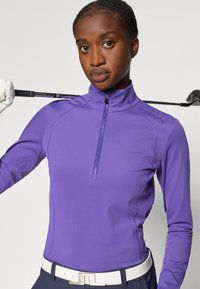 Haut de golf à manches longues violet avec un col montant, une fermeture éclair et une marque discrète. Le tissu semble lisse et extensible, améliorant le confort.