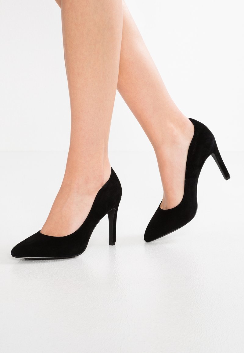 Pieds portant des escarpins en daim noir à bout pointu avec des talons aiguilles fins sur un sol blanc.