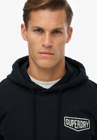 Sudadera con capucha azul marino con cordón ajustable y bolsillo frontal; presenta un logo blanco "SUPERDRY" bordado en el pecho. Textura de tela suave.