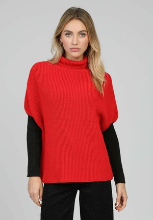 Pull rouge en maille avec col montant et manches courtes, manches longues noires en dessous, texture côtelée, associé à un pantalon noir.