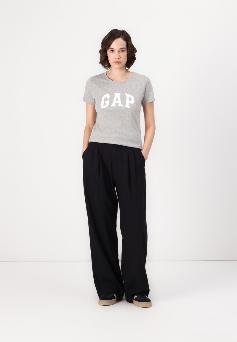 Tricou gri cu mânecă scurtă, cu logo-ul "GAP" în alb, asortat cu pantaloni largi negri și adidași negri cu tălpi de culoare deschisă.