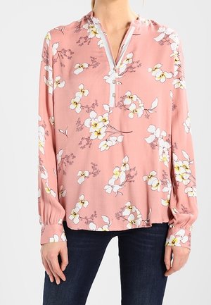 Blusa rosa con motivo floreale bianco, maniche lunghe, scollatura con bottoni e vestibilità comoda. Il tessuto sembra leggero e morbido.