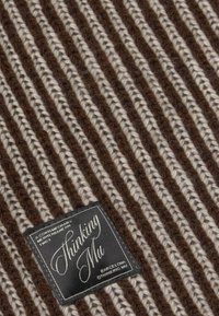Brown and beige striped knitted fabric with a black rectangular label reading "Thinking Mu, Barcelona" stitched on it.