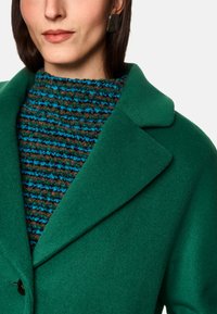 Grüner Wollmantel mit Reverskragen, kombiniert mit einem strukturierten, braun-blau gestreiften Strickoberteil. Verfügt über einen glatten Stoff und ein strukturiertes Design.