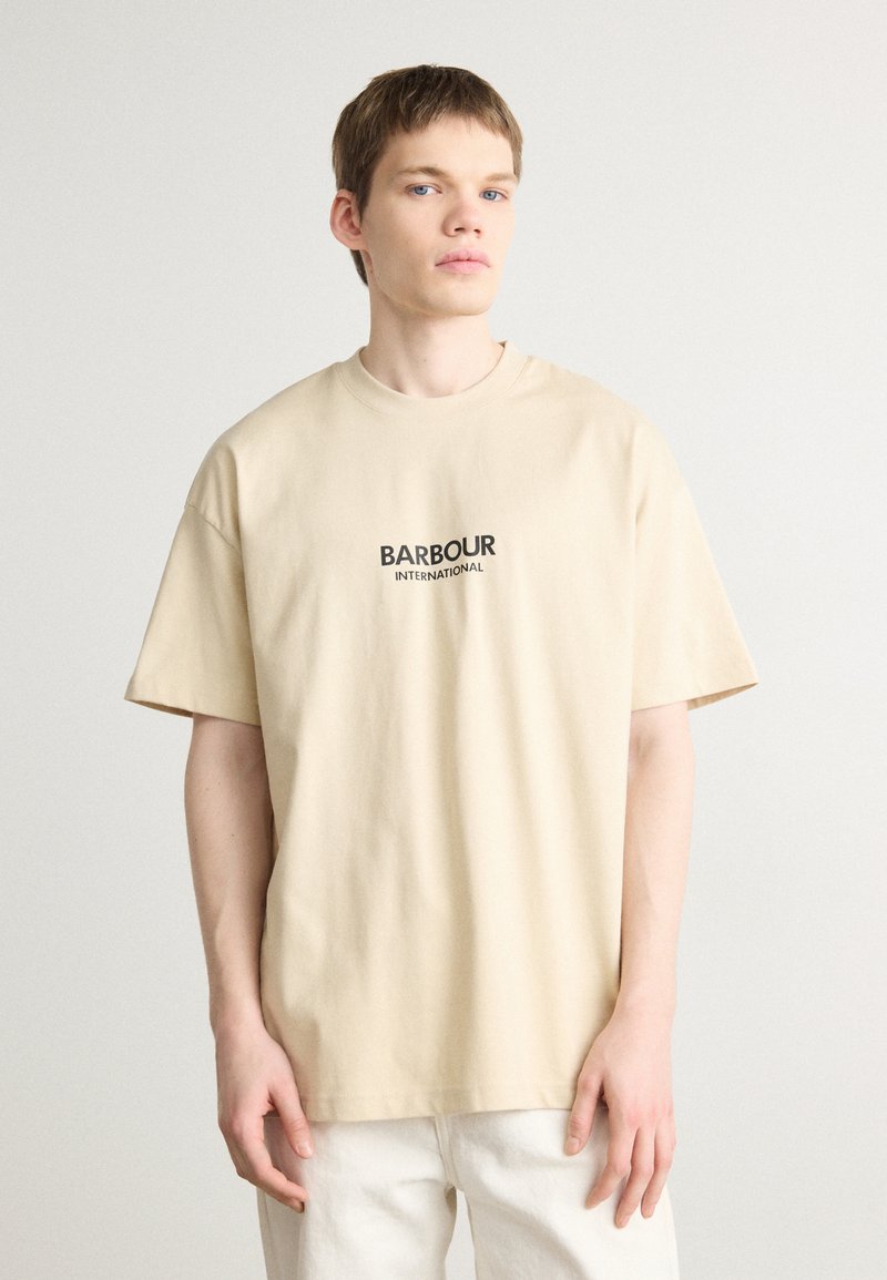 Beżowa bawełniana koszulka z krótkim rękawem, z nadrukowanym na czarno logo "BARBOUR INTERNATIONAL" na klatce piersiowej. Gładka tekstura.