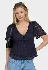 Jeune femme aux cheveux blonds portant un haut en dentelle bleu marine à manches bouffantes, un jean bleu clair, des boucles d'oreilles argentées et un sac à bandoulière noir.