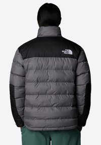 Grå puffjacka med en svart övre del och svarta accentdetaljer. Har en hög krage och en framträdande The North Face-logotyp på ryggen.