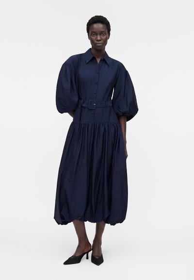 Γυναίκα στέκεται φορώντας ένα ναυτικό μπλε φόρεμα με κουμπιά, φουσκωτά μανίκια, ζώνη στη μέση και μαύρα ψηλοτάκουνα παπούτσια με μυτερή μύτη.