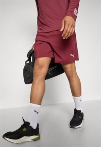 Weinrote Sportshorts kombiniert mit einem weinroten Langarmshirt, schwarzen Turnschuhen mit grünen Akzenten und weißen Socken. Eine schwarze Sporttasche wird gehalten.