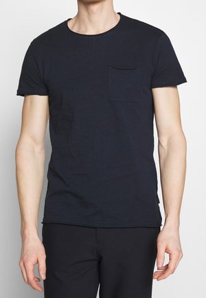 Mann trägt ein einfaches schwarzes T-Shirt mit kurzen Ärmeln und einer Brusttasche, kombiniert mit schwarzen Hosen, steht vor einem schlichten Hintergrund.