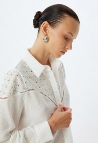Camicia bianca con bottoni, decorata con dettagli ricamati a paisley, in tessuto testurizzato e colletto classico, abbinata a orecchini a cerchio argentati.
