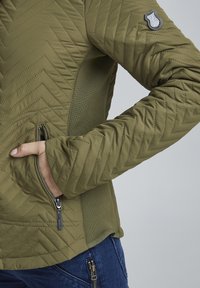 Veste matelassée vert olive avec motif en chevron, poches zippées et panneaux latéraux texturés. Présente une patch logo sur le bras.