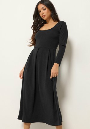 Femme portant une longue robe noire à manches longues et un décolleté arrondi, posant devant un fond clair uni.